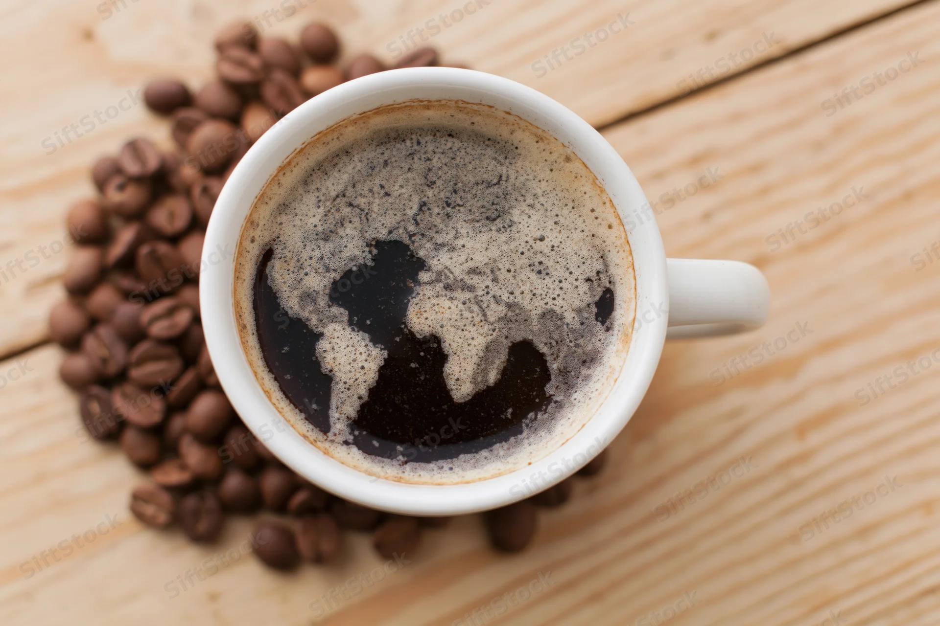 White ceramic mug filled with black coffee showing foam map of continents on wooden table surrounded by roasted beans