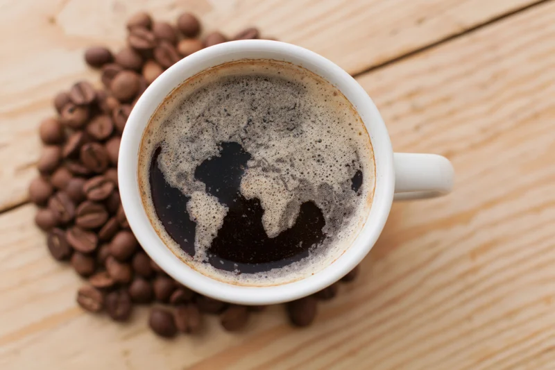 White ceramic mug filled with black coffee showing foam map of continents on wooden table surrounded by roasted beans