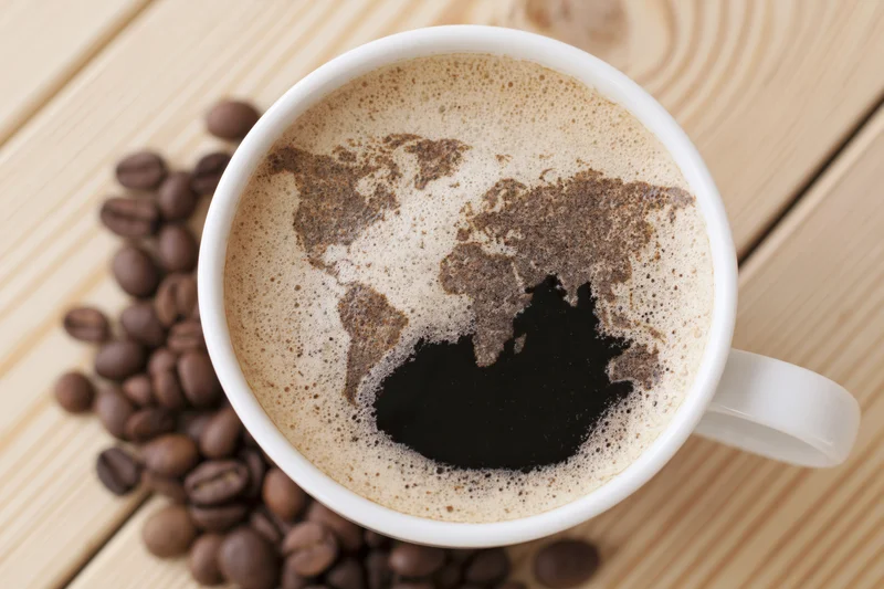 White ceramic mug filled with coffee featuring world map in foam on wooden table with scattered coffee beans around