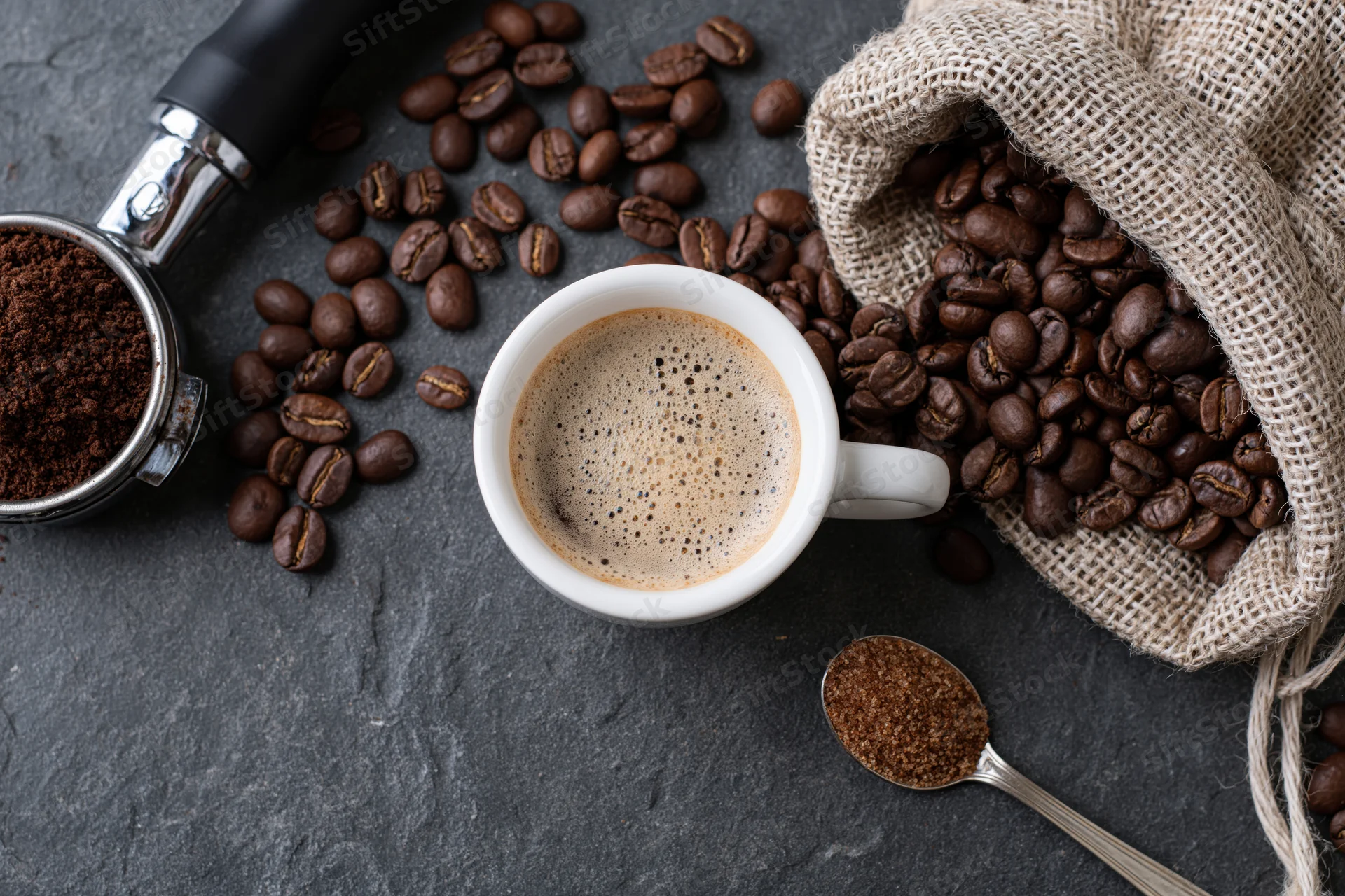 White espresso cup with frothy coffee in overhead flat lay surrounded by beans, grinder, and burlap sack on dark stone surface
