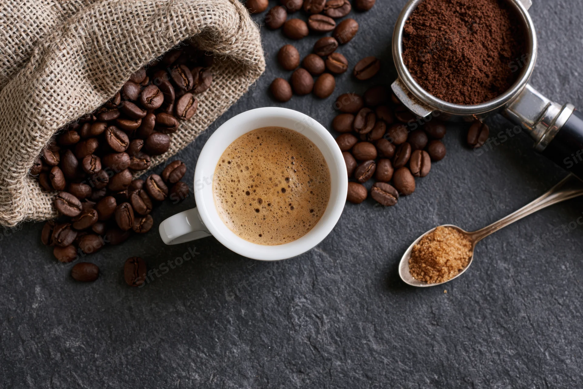 Overhead flat lay of a white espresso cup filled with frothy coffee surrounded by beans, grinder, and sugar on dark stone surface