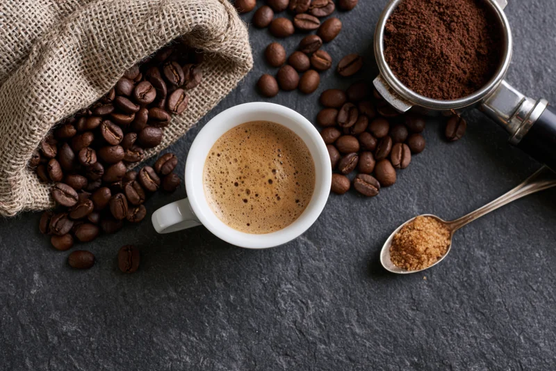 Overhead flat lay of a white espresso cup filled with frothy coffee surrounded by beans, grinder, and sugar on dark stone surface