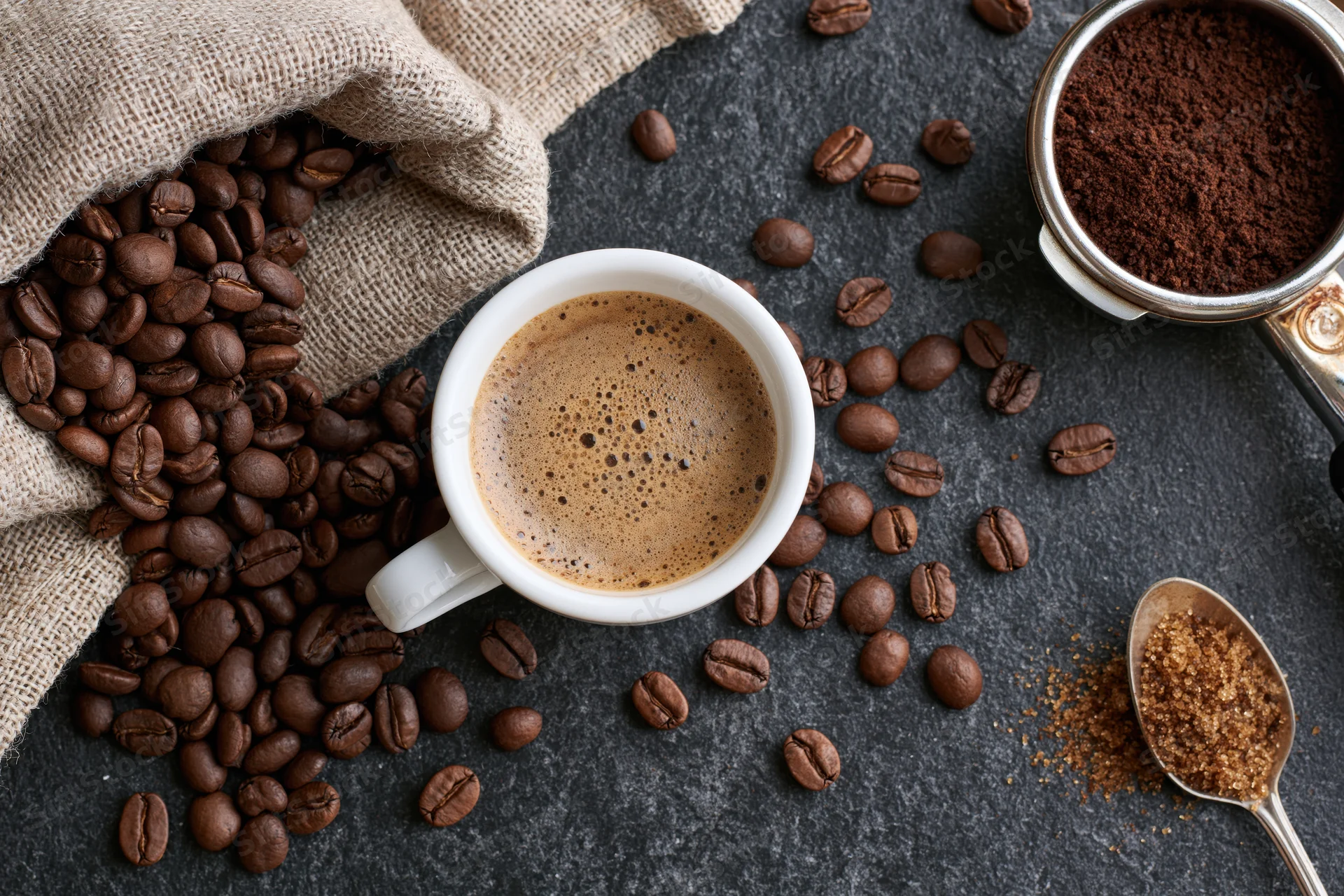 White espresso cup filled with frothy coffee in overhead flat lay with scattered beans and sugar on dark stone surface