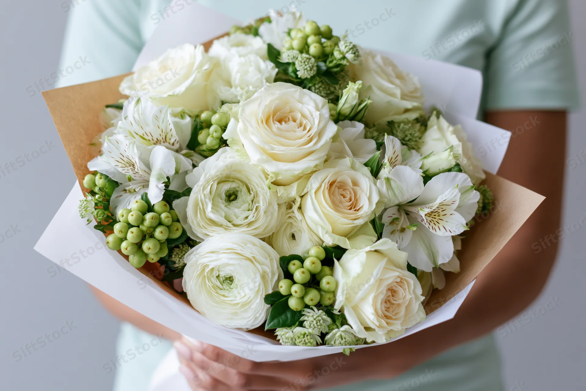 Person holding a large bouquet of white roses, ranunculus, and alstroemeria with green berries and foliage wrapped in paper