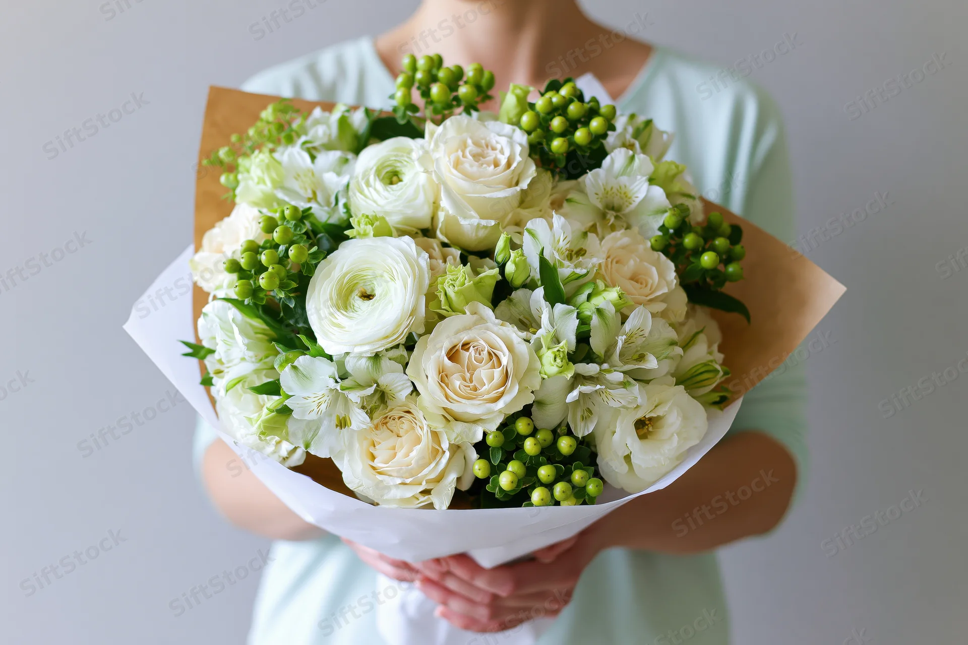 Woman holding a large bouquet of white roses, ranunculus and alstroemeria with green berries and foliage wrapped in paper