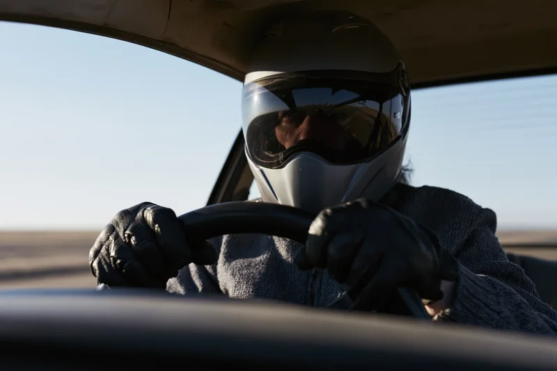 Driver in full-face motorcycle helmet gripping steering wheel inside car on open road