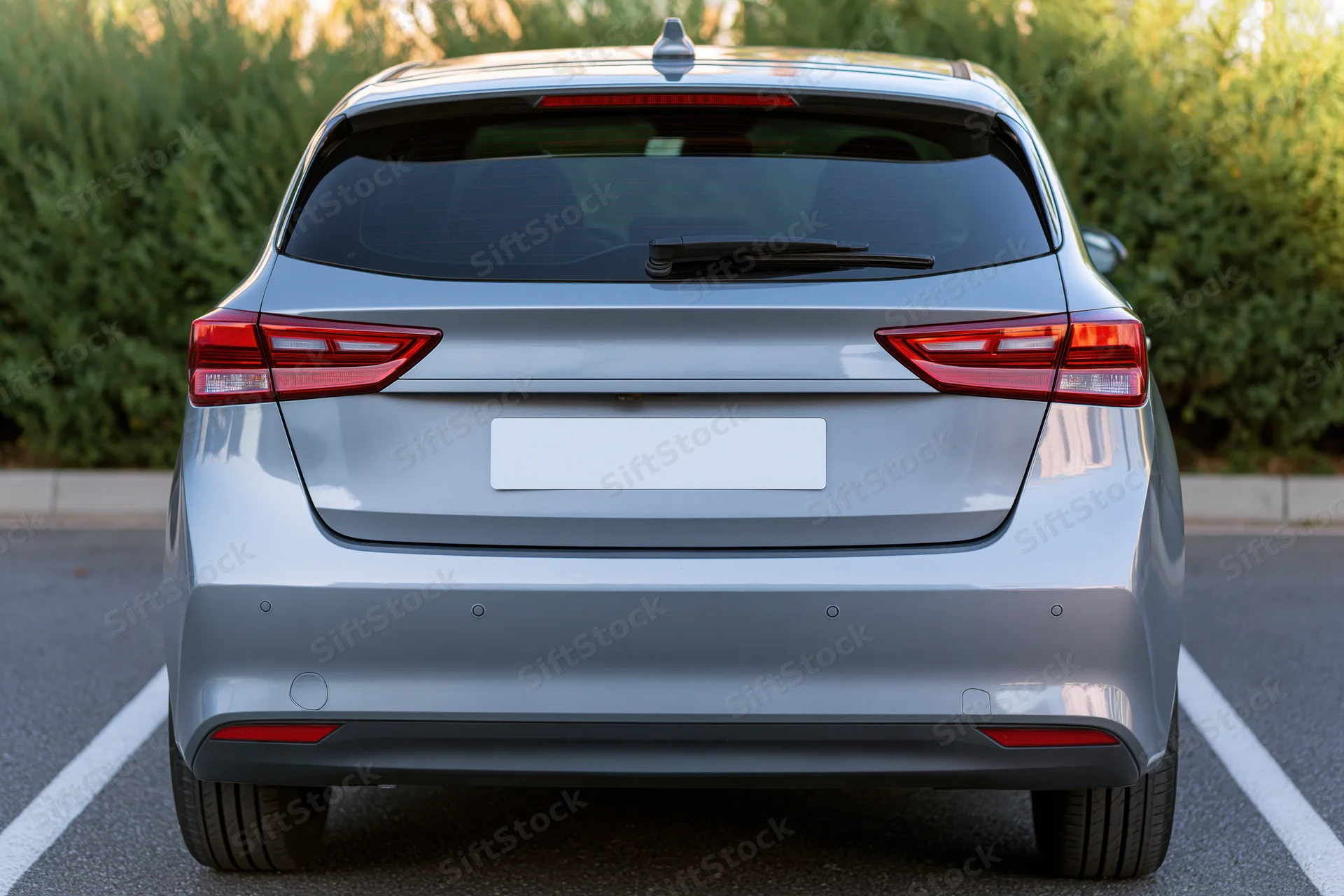 Rear view of a silver compact hatchback parked in an outdoor lot with white parking lines and green hedge background