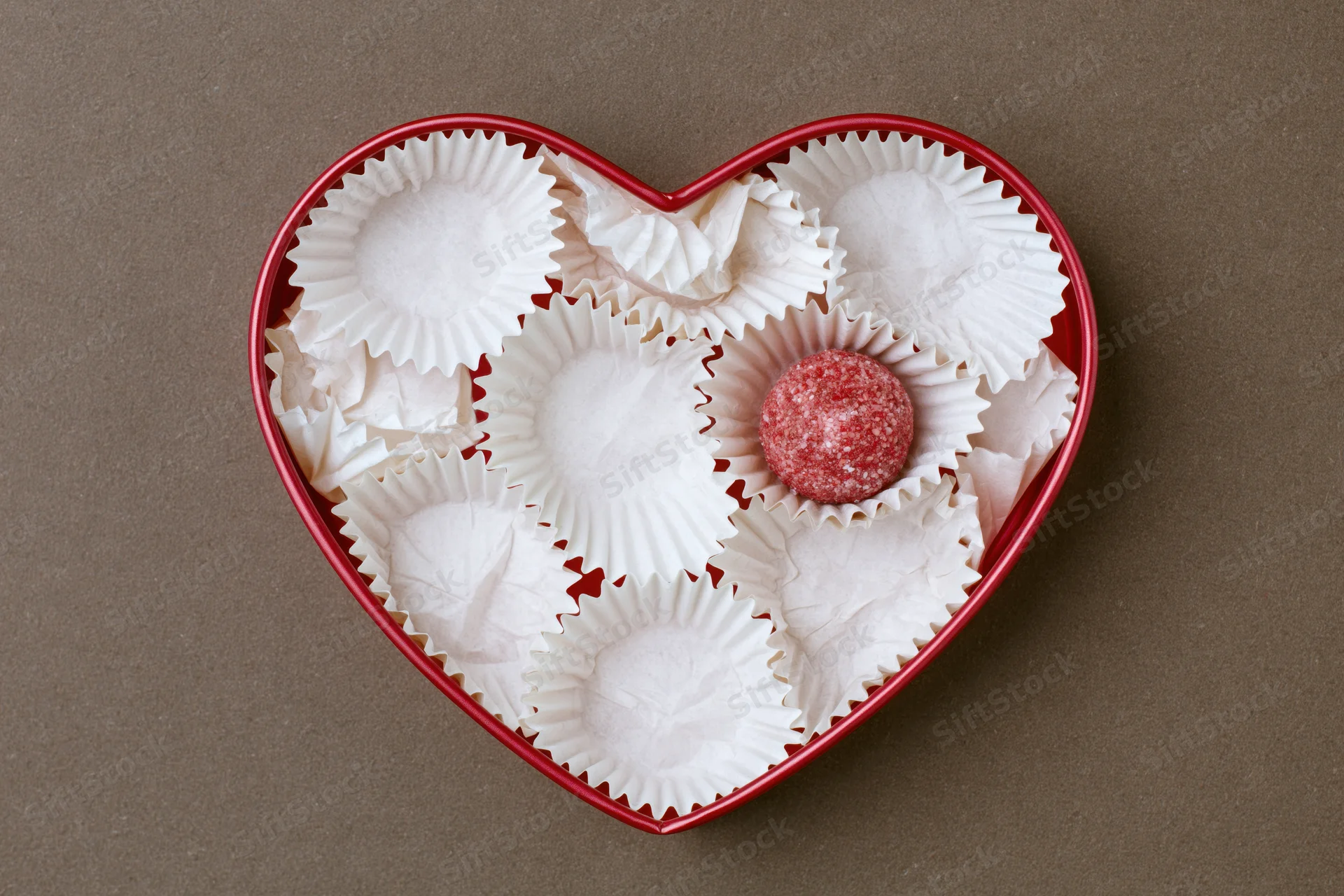 Red heart-shaped gift box filled with white paper candy wrappers and one red chocolate candy in center on brown surface