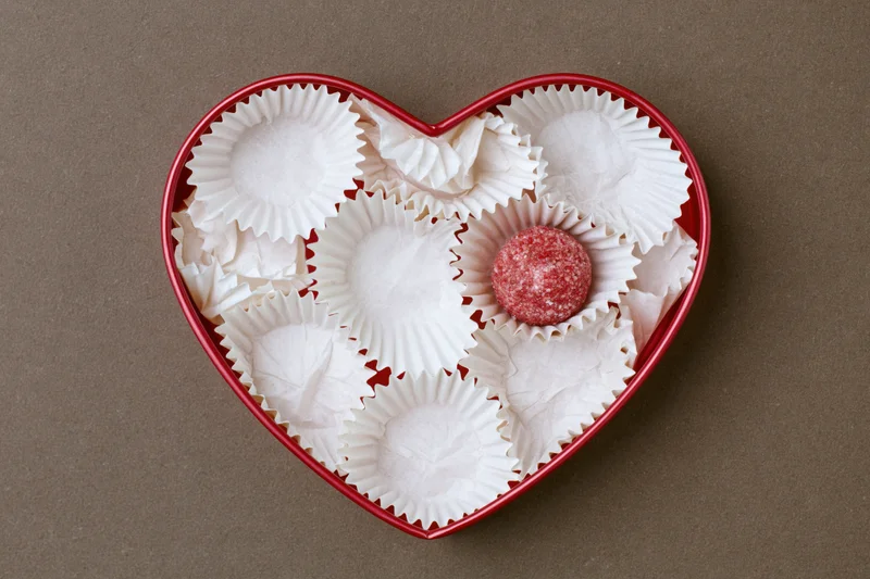 Red heart-shaped gift box filled with white paper candy wrappers and one red chocolate candy in center on brown surface