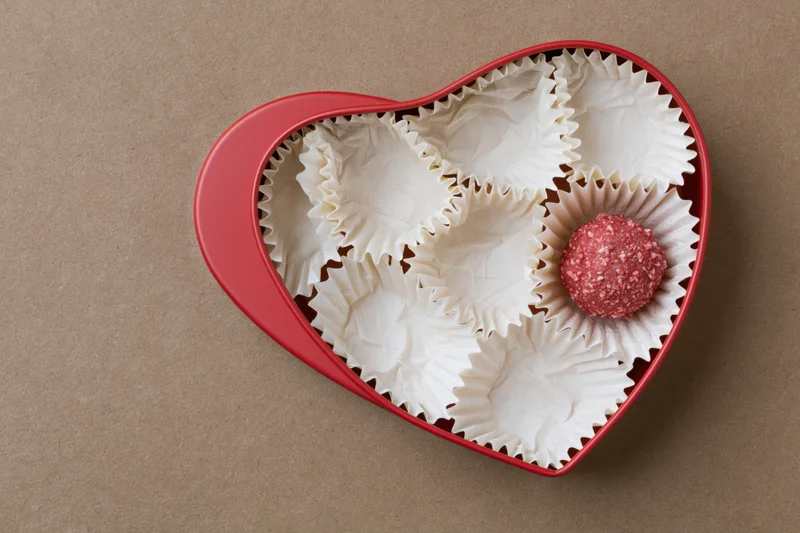 Red heart-shaped box with white paper candy wrappers and one chocolate truffle inside on brown surface