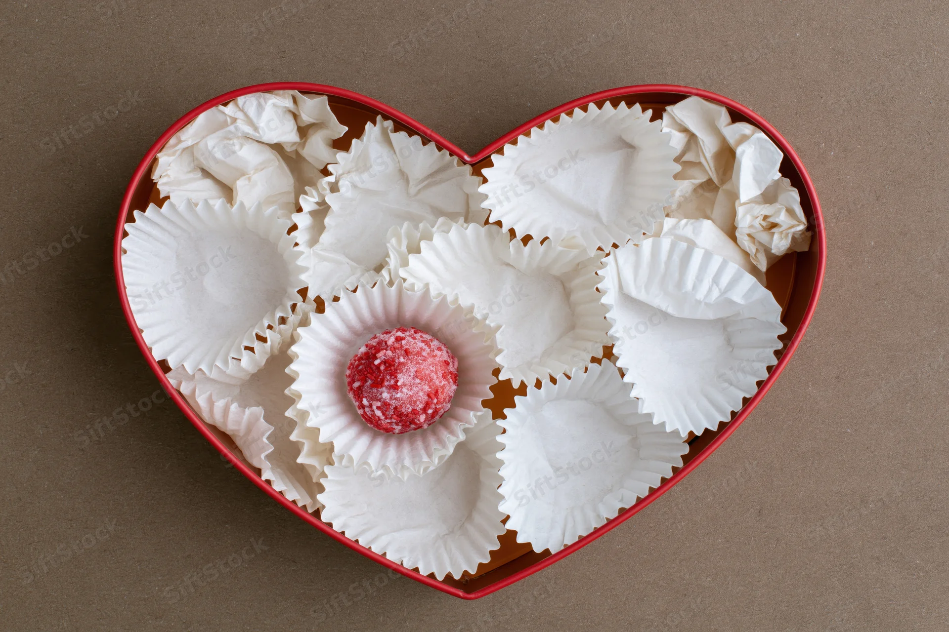 Red heart-shaped gift box containing a single red candy in a white paper wrapper surrounded by empty wrappers on a brown surface