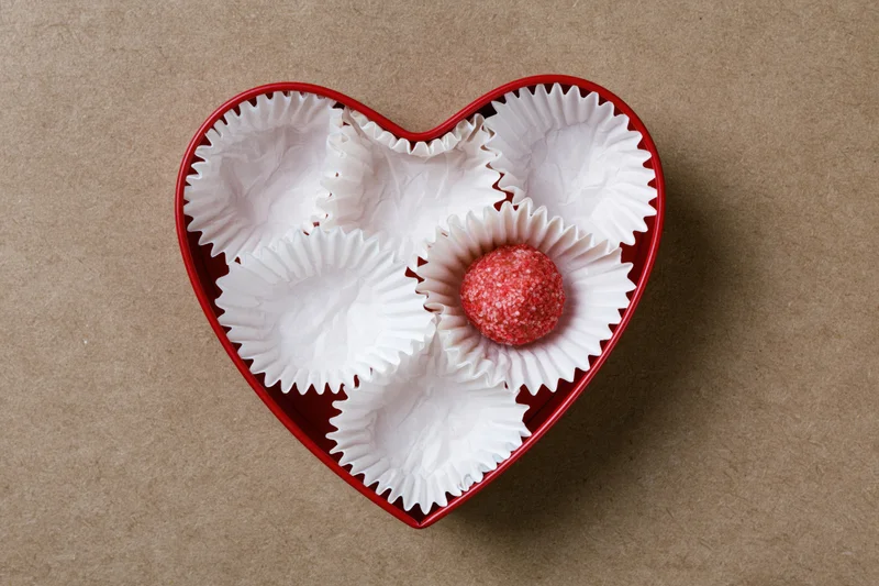 Red heart-shaped box holding white paper candy liners with one red chocolate truffle inside on brown surface