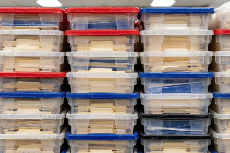 Rows of clear plastic storage bins with red blue and black lids stacked vertically in an office filing system