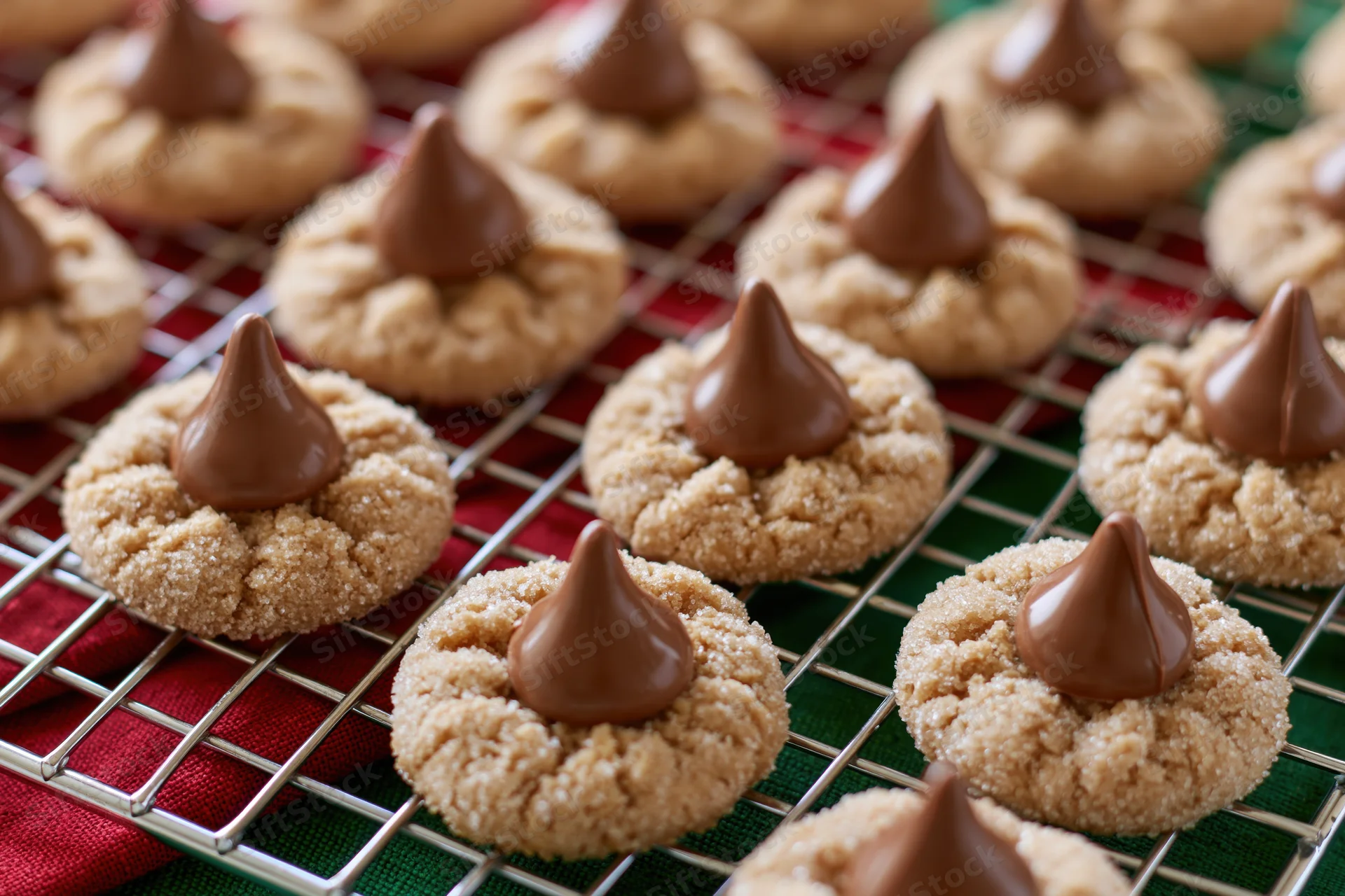 Rows of peanut butter blossom cookies topped with chocolate kisses cooling on a wire rack over red and green cloth