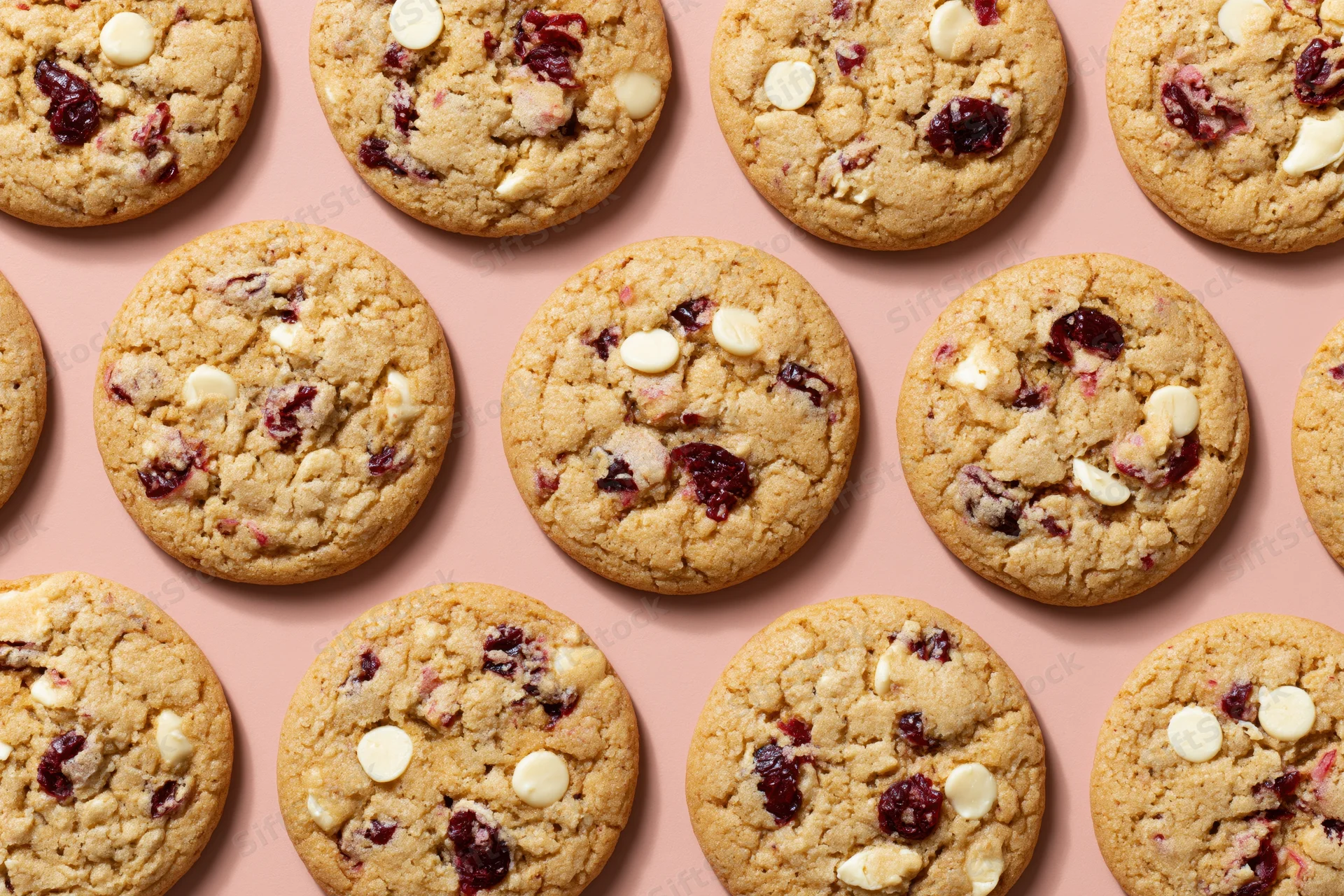 Rows of round cranberry and white chocolate chunk cookies arranged in a grid on a pink surface