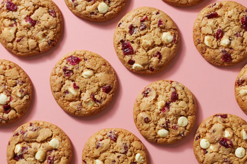 Round cranberry and white chocolate chunk cookies arranged in rows on a pink surface