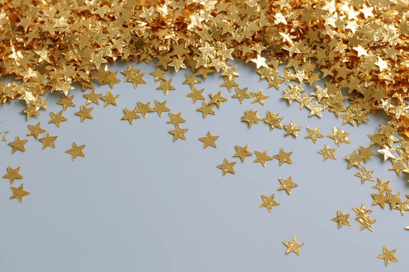 Gold metallic star-shaped confetti scattered across a light blue surface with copyspace