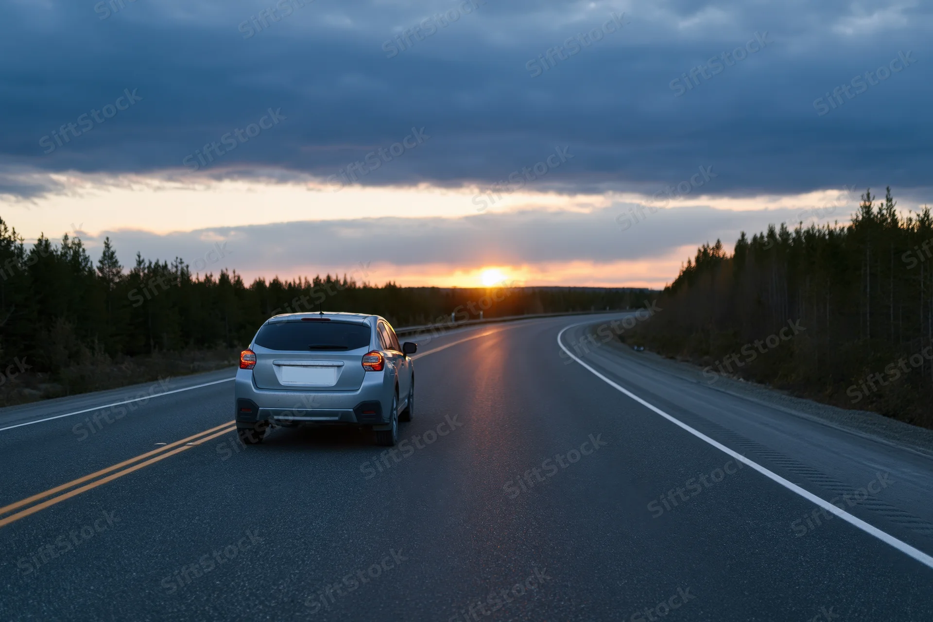 Silver suv driving away on a winding two-lane highway through a forest at sunset