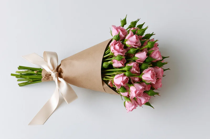 Small bouquet of pink spray roses and green buds wrapped in kraft paper with cream ribbon on white background