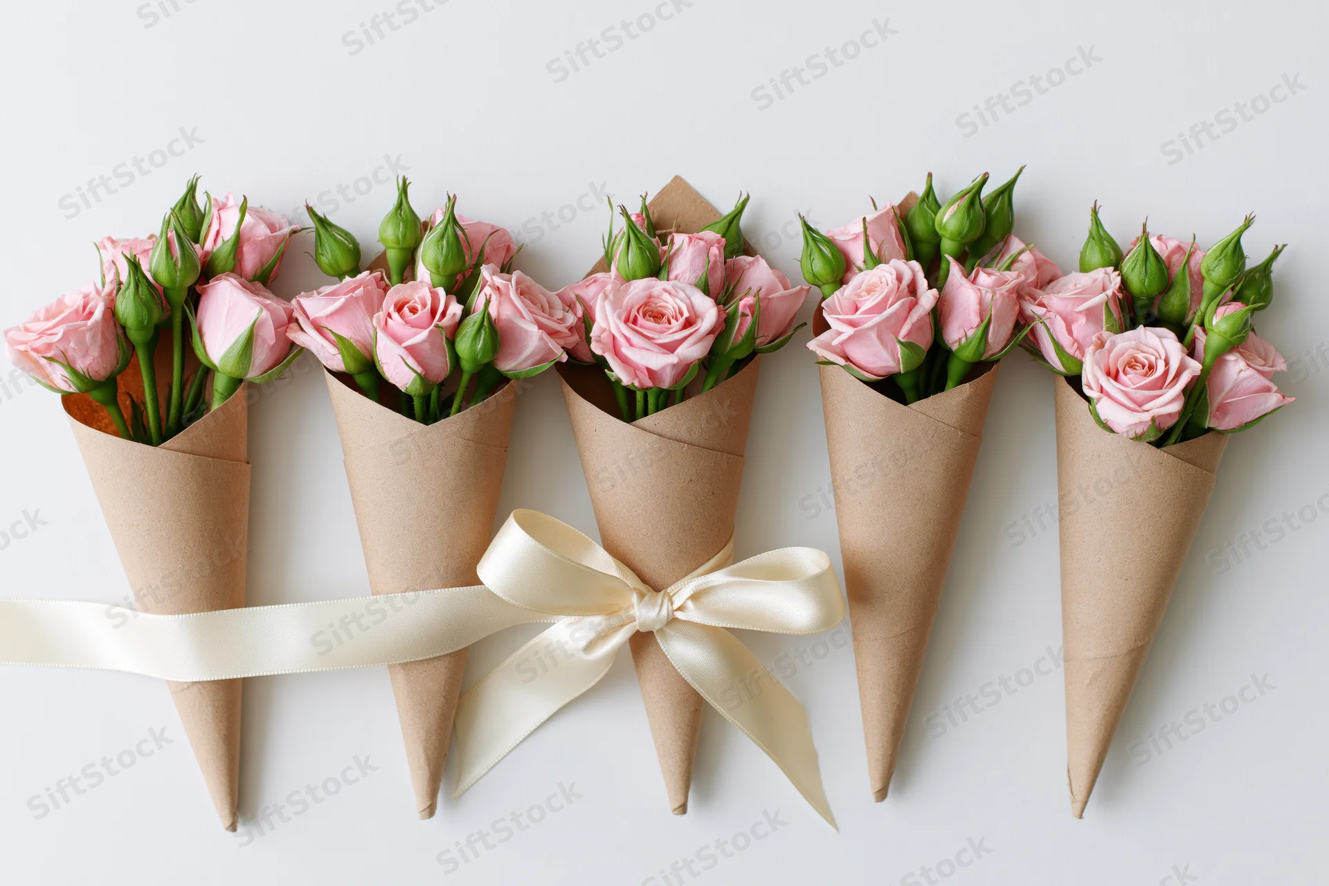 Small bouquets of pink spray roses and green buds wrapped in kraft paper cones arranged in a row with a cream ribbon on white background
