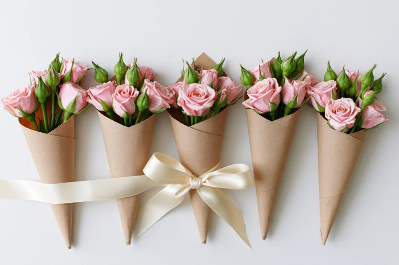 Small bouquets of pink spray roses and green buds wrapped in kraft paper cones arranged in a row with a cream ribbon on white background