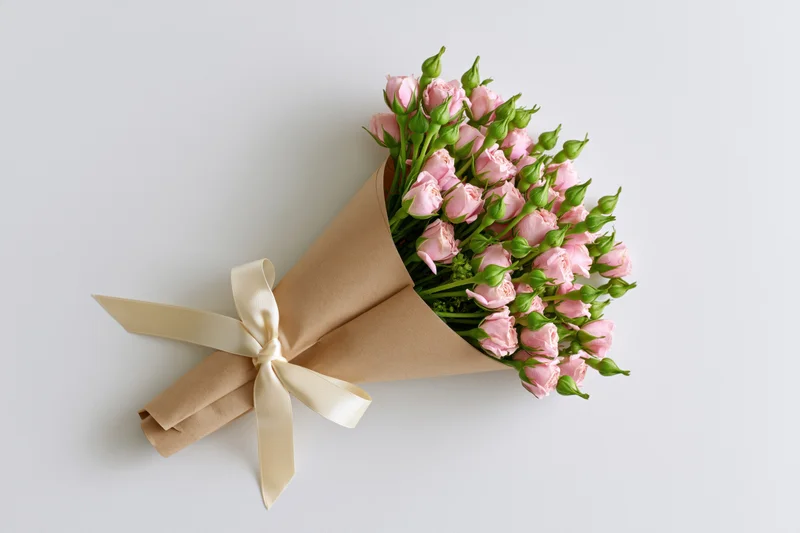 Small bouquet of pink spray roses and green buds wrapped in kraft paper with cream ribbon on white background
