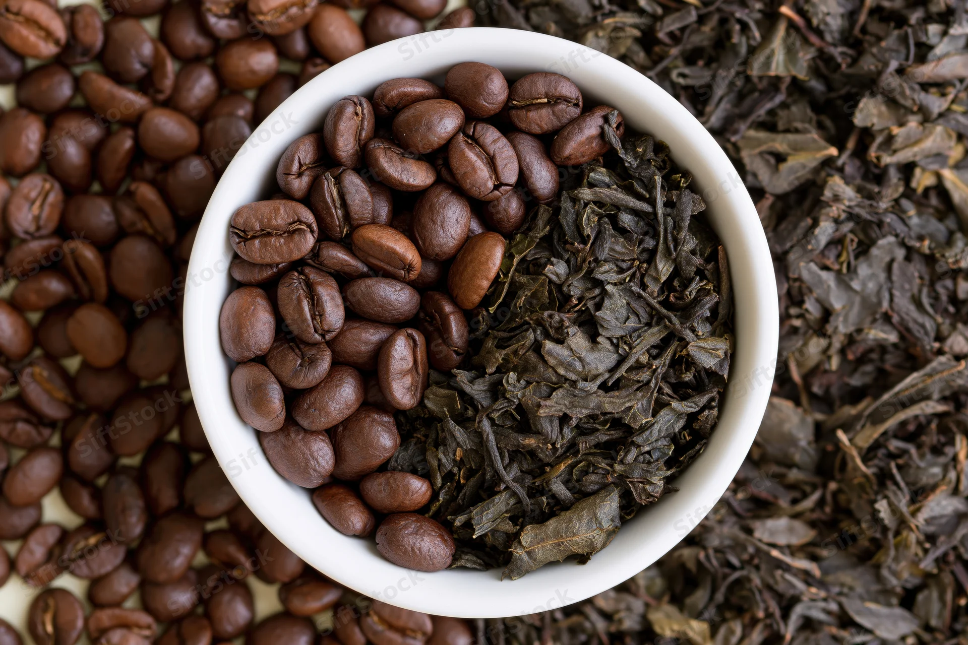 White ceramic bowl filled half with roasted coffee beans and half with dried tea leaves, surrounded by scattered beans and leaves