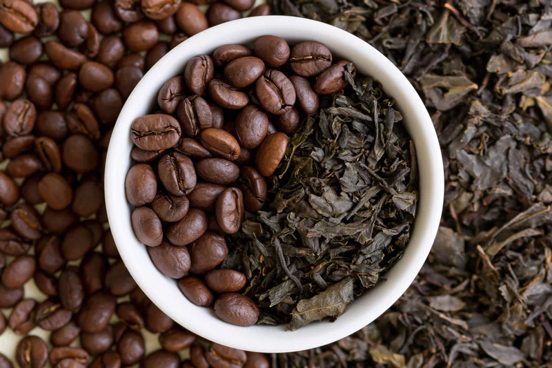 White ceramic bowl filled half with roasted coffee beans and half with dried tea leaves, surrounded by scattered beans and leaves
