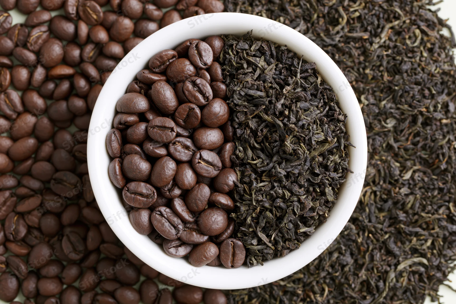 White ceramic bowl split with roasted coffee beans and dark tea leaves arranged in half-and-half pattern on scattered beans and leaves background