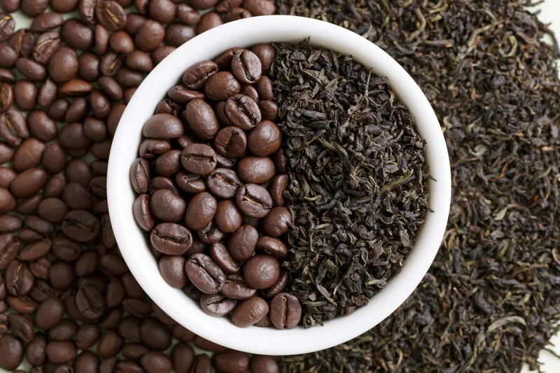White ceramic bowl split with roasted coffee beans and dark tea leaves arranged in half-and-half pattern on scattered beans and leaves background