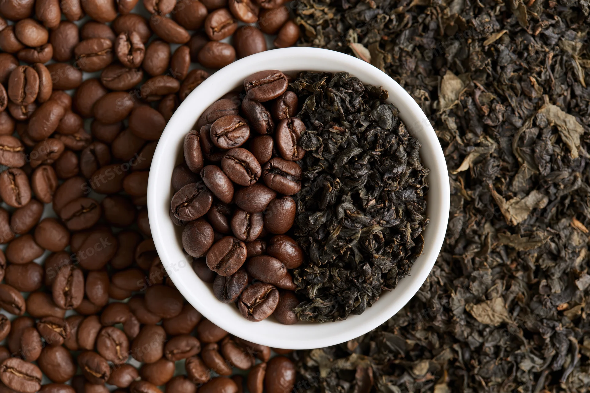 Small white ceramic bowl split with roasted coffee beans and dark dried tea leaves arranged on a surface of scattered beans and leaves