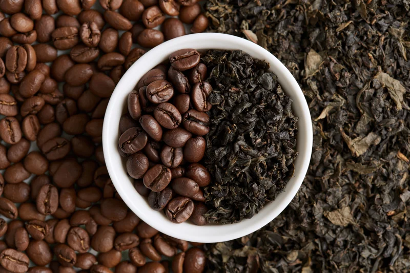 Small white ceramic bowl split with roasted coffee beans and dark dried tea leaves arranged on a surface of scattered beans and leaves