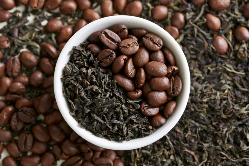 White ceramic bowl filled with roasted coffee beans and dried tea leaves, arranged on a scattered bed of beans and leaves