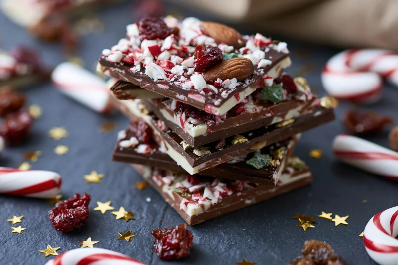 Stack of layered milk and dark chocolate bark with crushed candy canes, almonds, and dried cranberries on dark surface surrounded by festive decorations