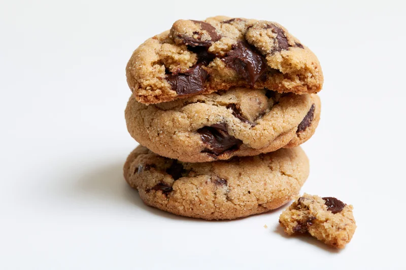 Stack of three chocolate chip cookies on white background with one broken piece beside