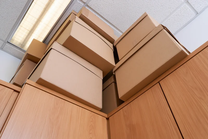 Stacked cardboard moving boxes piled high on top of a pine wood cabinet in an office interior