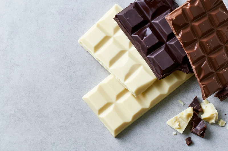 Three chocolate bars dark white and milk arranged overlapping on gray surface with broken pieces scattered nearby