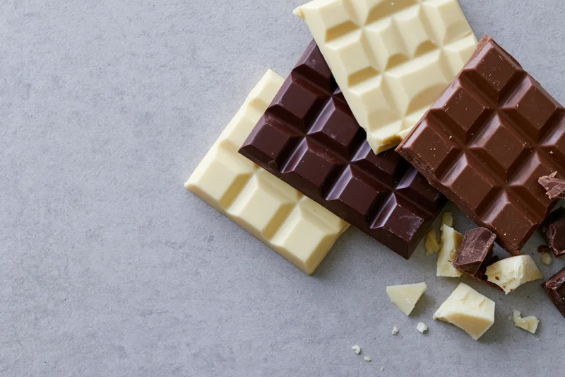 Overlapping dark, milk and white chocolate bars with broken pieces on gray surface