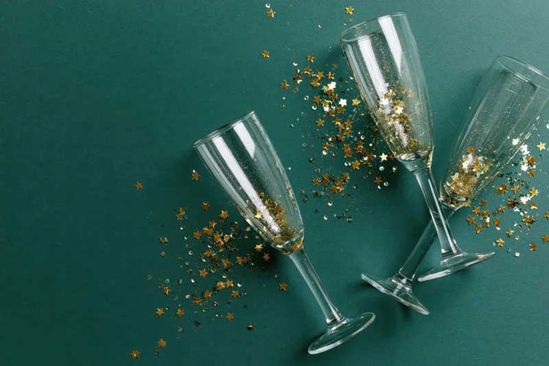 Three empty champagne flutes arranged on a flat deep green background with scattered gold star confetti