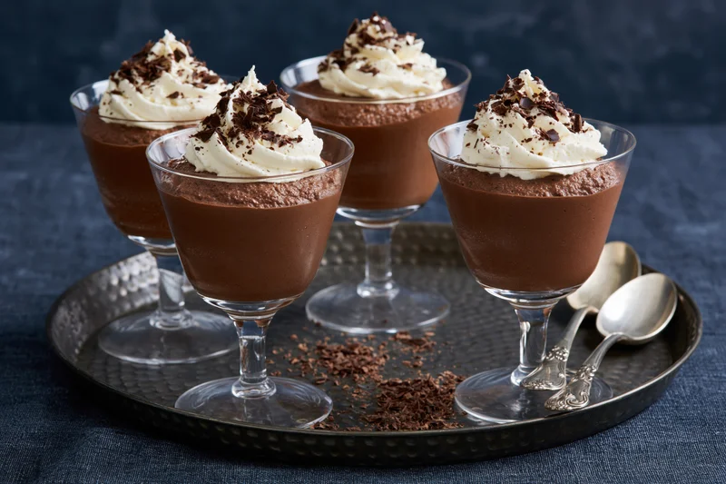 Chocolate mousse desserts in stemmed glasses topped with whipped cream and chocolate shavings on a silver tray with spoons