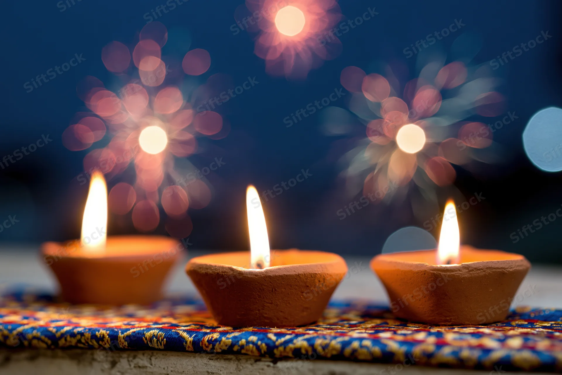 Three lit clay diya oil lamps arranged on a patterned fabric with bokeh fireworks in the background
