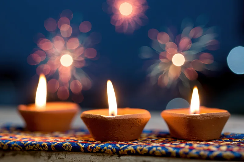 Three lit clay diya oil lamps arranged on a patterned fabric with bokeh fireworks in the background