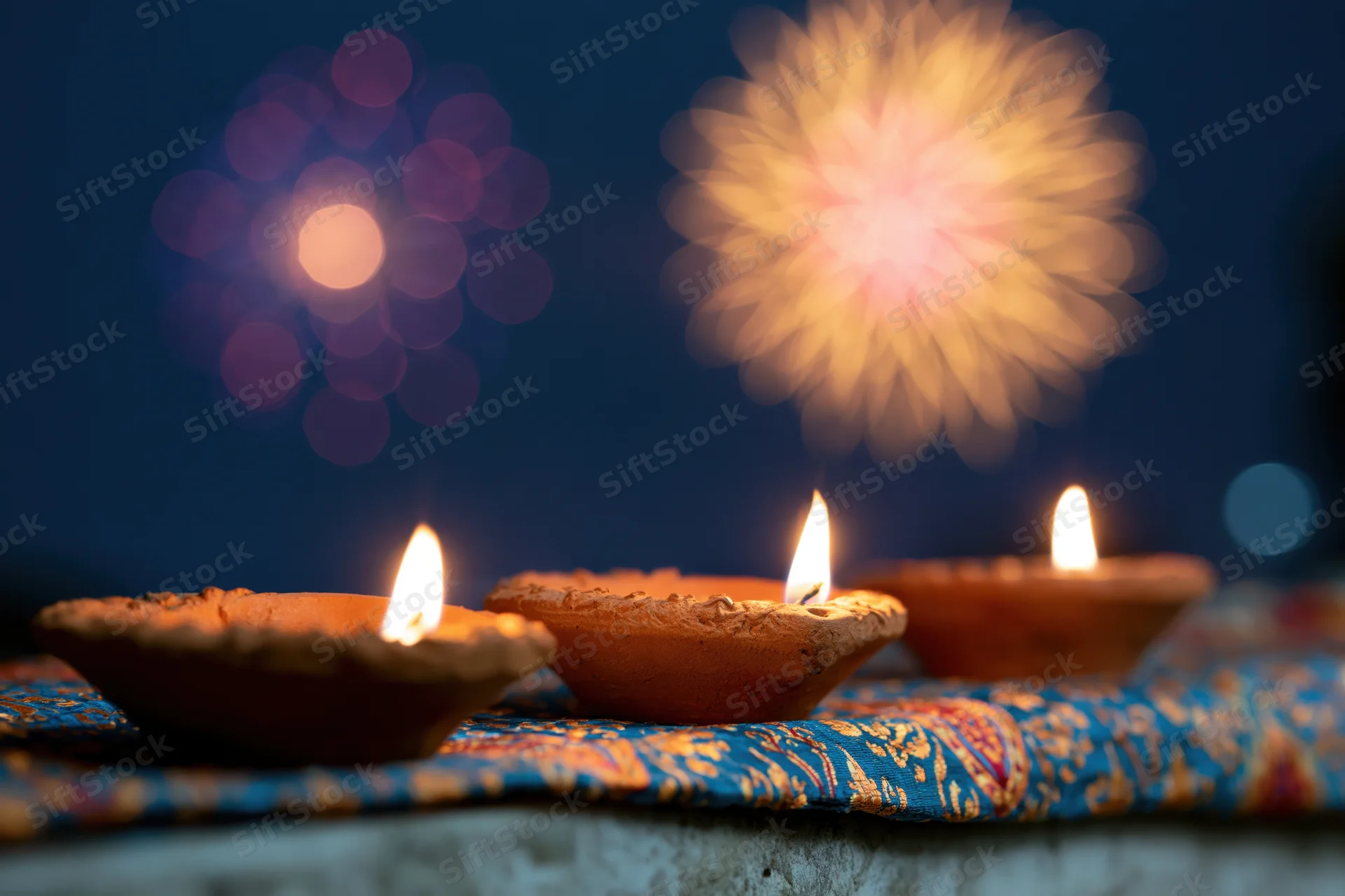 Three lit clay diya oil lamps arranged in a row on a patterned fabric at night with bokeh fireworks in the background