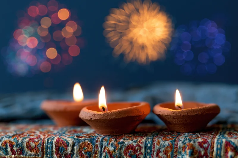Three lit clay diya oil lamps arranged on a patterned fabric with blurred fireworks in the background