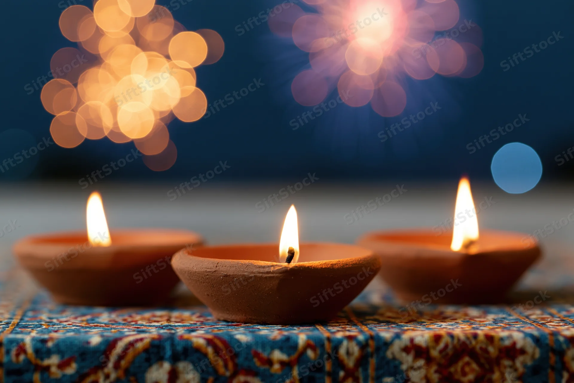 Three lit clay diya oil lamps arranged in a row on a decorative fabric surface with bokeh fireworks in the background