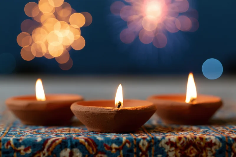Three lit clay diya oil lamps arranged in a row on a decorative fabric surface with bokeh fireworks in the background