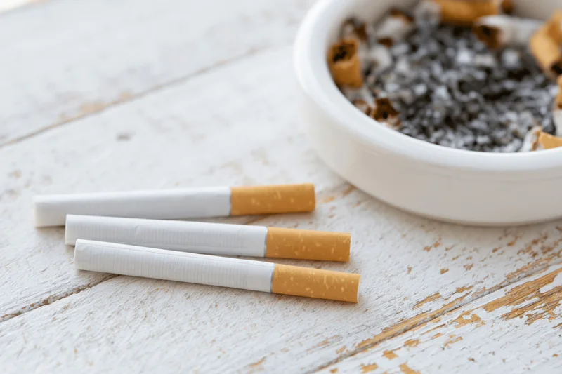 Three unlit cigarettes lying flat on a worn white wooden surface next to an ashtray filled with ash and cigarette butts