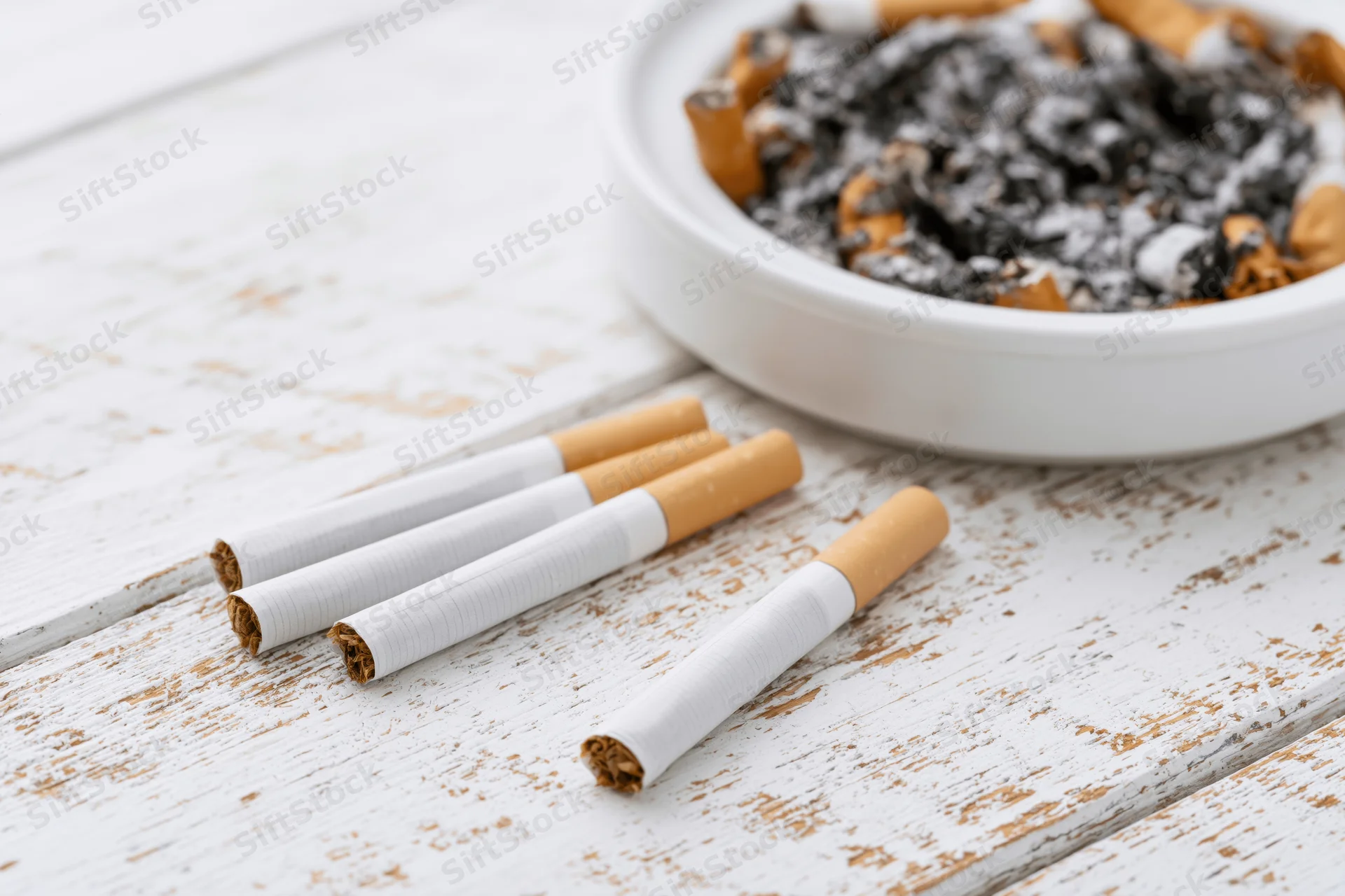 Unlit cigarettes lying flat on a worn white wooden surface with an ashtray containing cigarette butts and ash nearby