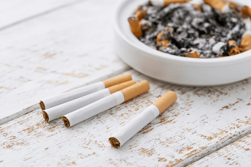 Unlit cigarettes lying flat on a worn white wooden surface with an ashtray containing cigarette butts and ash nearby