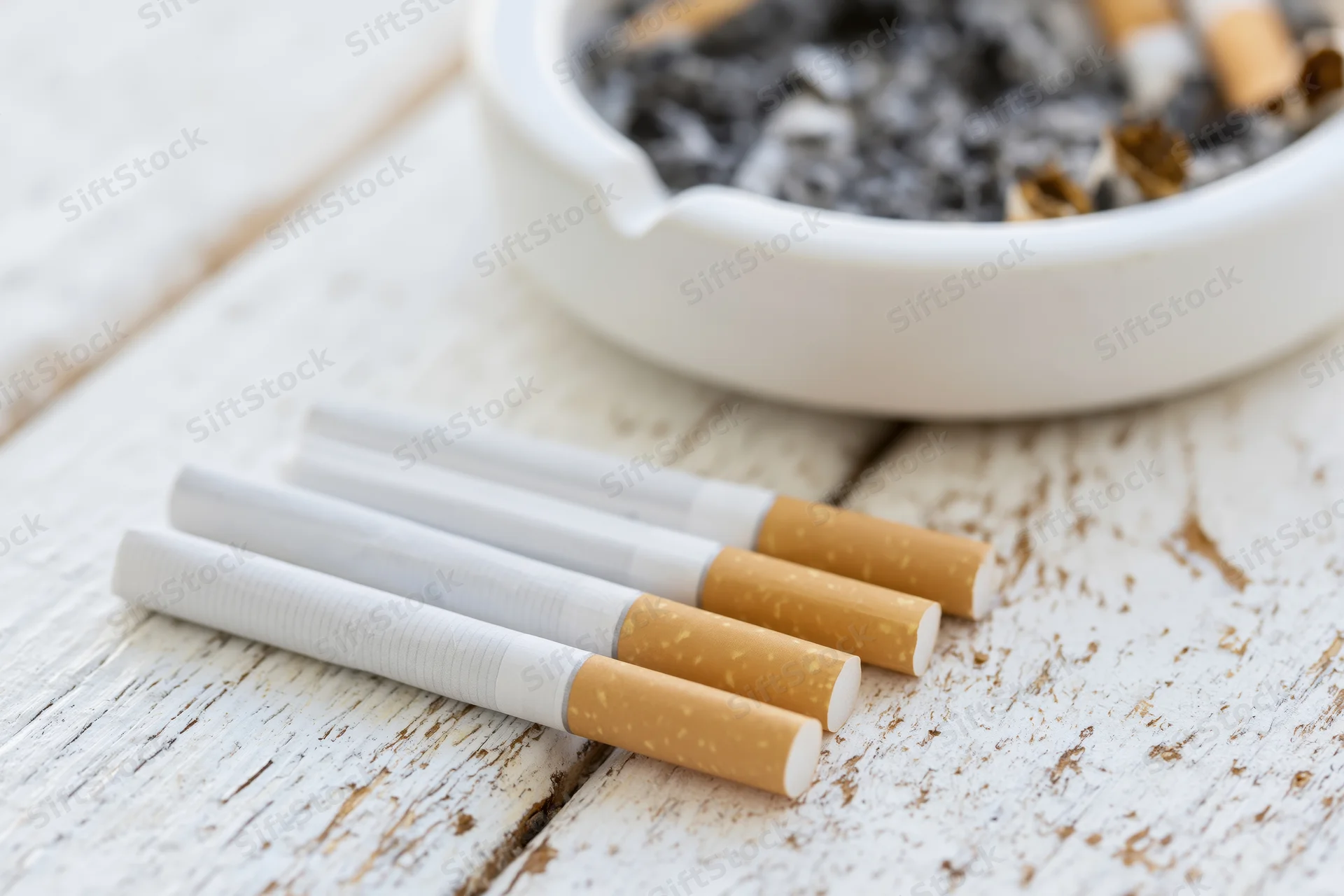 Three unlit cigarettes lying flat on a worn white wooden surface with an ashtray in the background